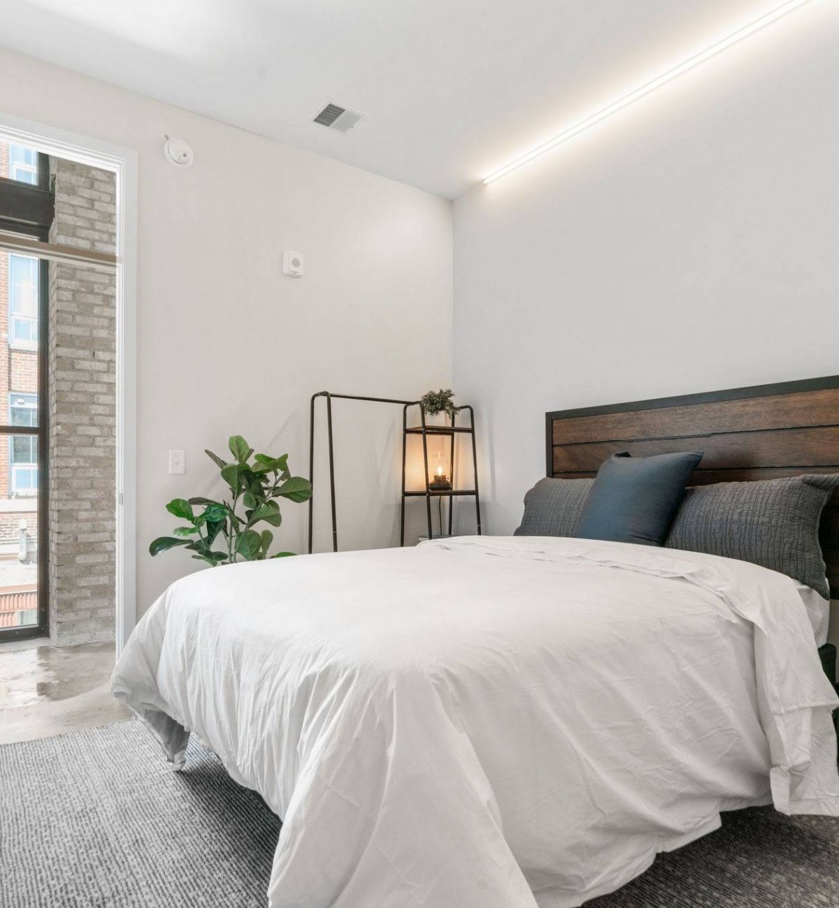A bright bedroom in Factory Lofts with a white bedspread, wooden headboard, and large window overlooking the brick building outside.